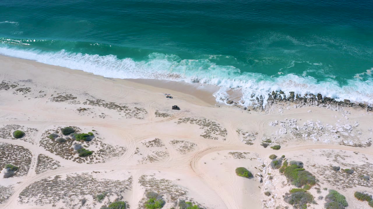 Overhead view in Los Cabos, Mexico of teal surf rolling toward pale sand, dark shoreline rocks, scrub patches, and no visible people.