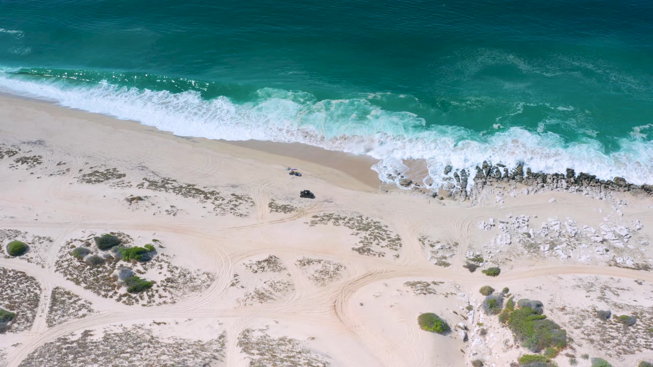 Overhead Los Cabos, Mexico shoreline frame with deeper teal water at the top, a band of white surf, pale open sand, scattered rocks, and no visible people.