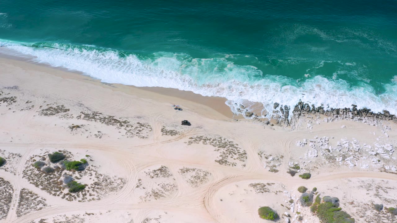 Los Cabos, Mexico aerial view with surf pressing toward a rocky beach, pale sand below, scrub clusters near the lower edge, and no visible people.