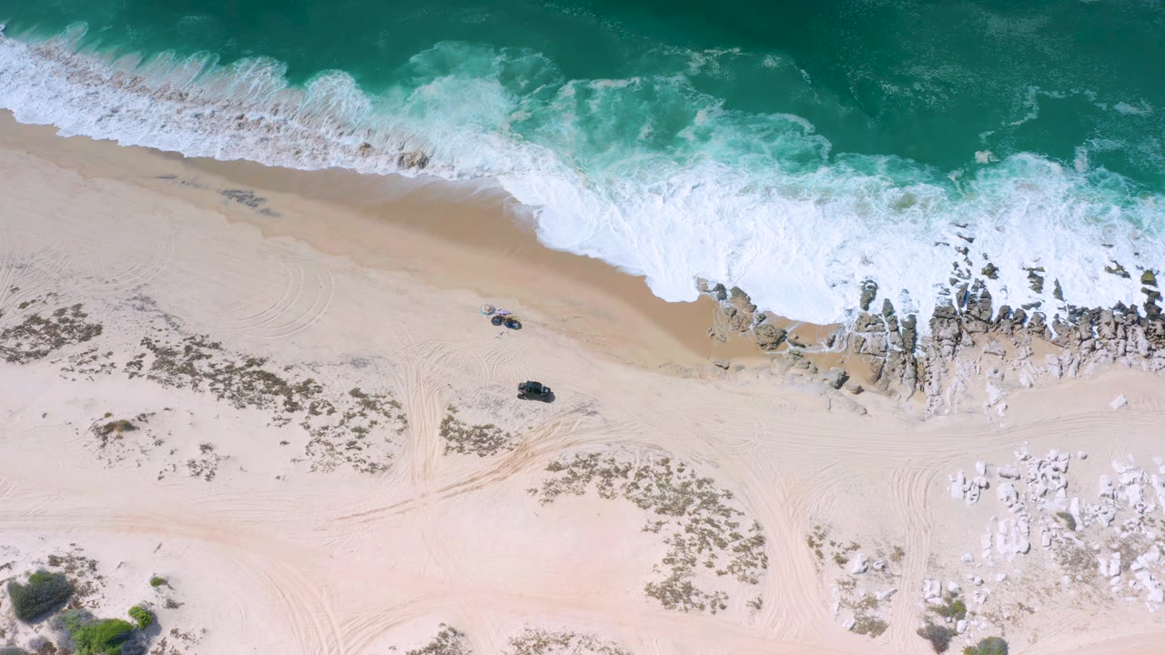 Aerial Los Cabos shoreline frame with the wave band pushed farther across the upper beach, two small dark beach objects, rocks near the waterline, and no visible people.