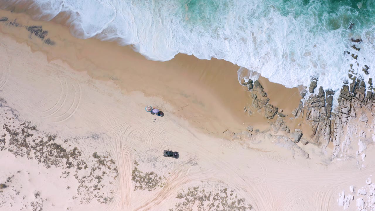 Los Cabos overhead beach view with white foam pushing toward tan sand, a rocky edge on the right, curved beach tracks, and two small objects on the open sand with no visible people.
