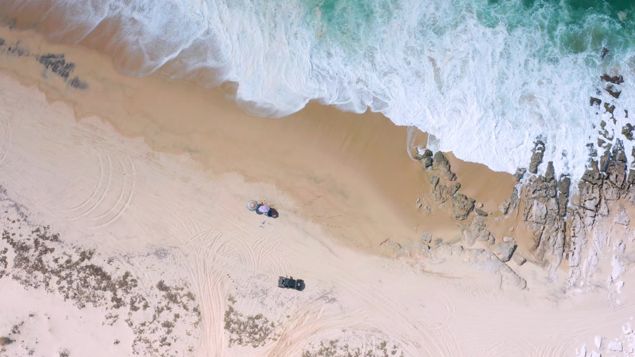 Direct overhead Los Cabos frame with the surf line farther across the top edge, tan beach below, jagged rocks on the right, and small dark and light objects on the sand with no visible people.