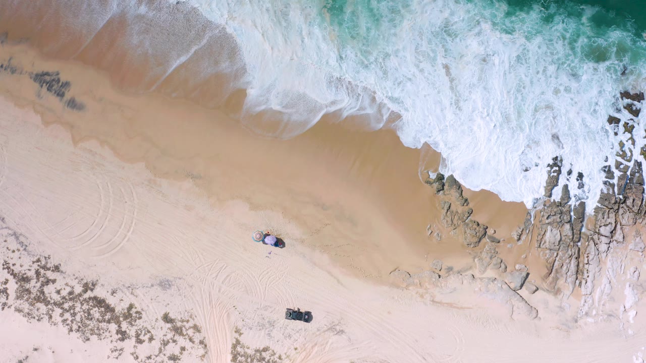 Los Cabos aerial view where whitewater covers more of the upper shoreline, tan wet sand sits below it, rocks run along the right side, and faint vehicle-like tracks curve through the sand with no visible people.