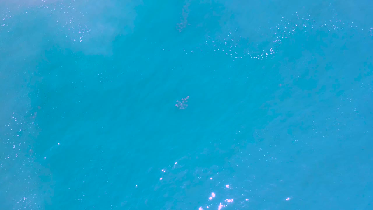 Aerial frame of light blue water in Los Cabos, Mexico, with faint foam patches across the top, subtle wave texture, reflected highlights near the lower left, and no visible people.