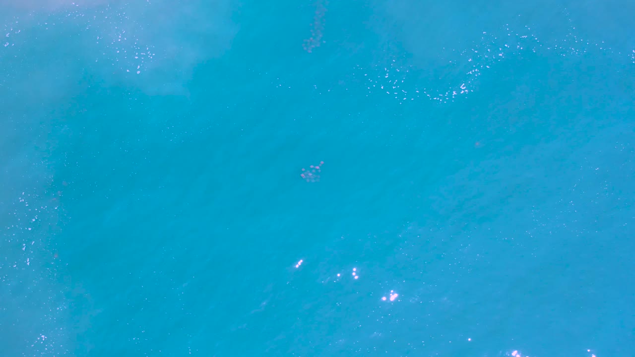 Overhead Los Cabos ocean frame with turquoise water, soft underwater color variation, small white foam clusters near the top, shimmer spots toward the right, and no visible people.