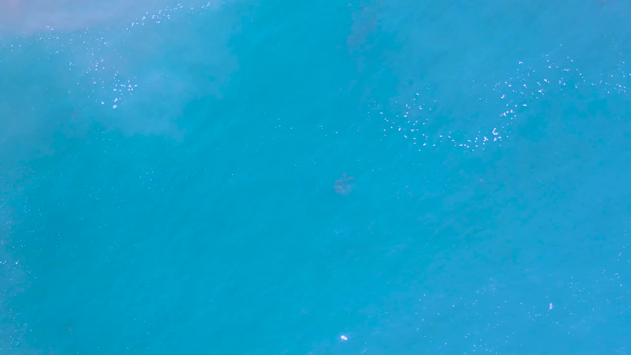 Aerial turquoise water in Los Cabos, Mexico, with a soft foam cluster near the top and scattered reflective points, no visible people.