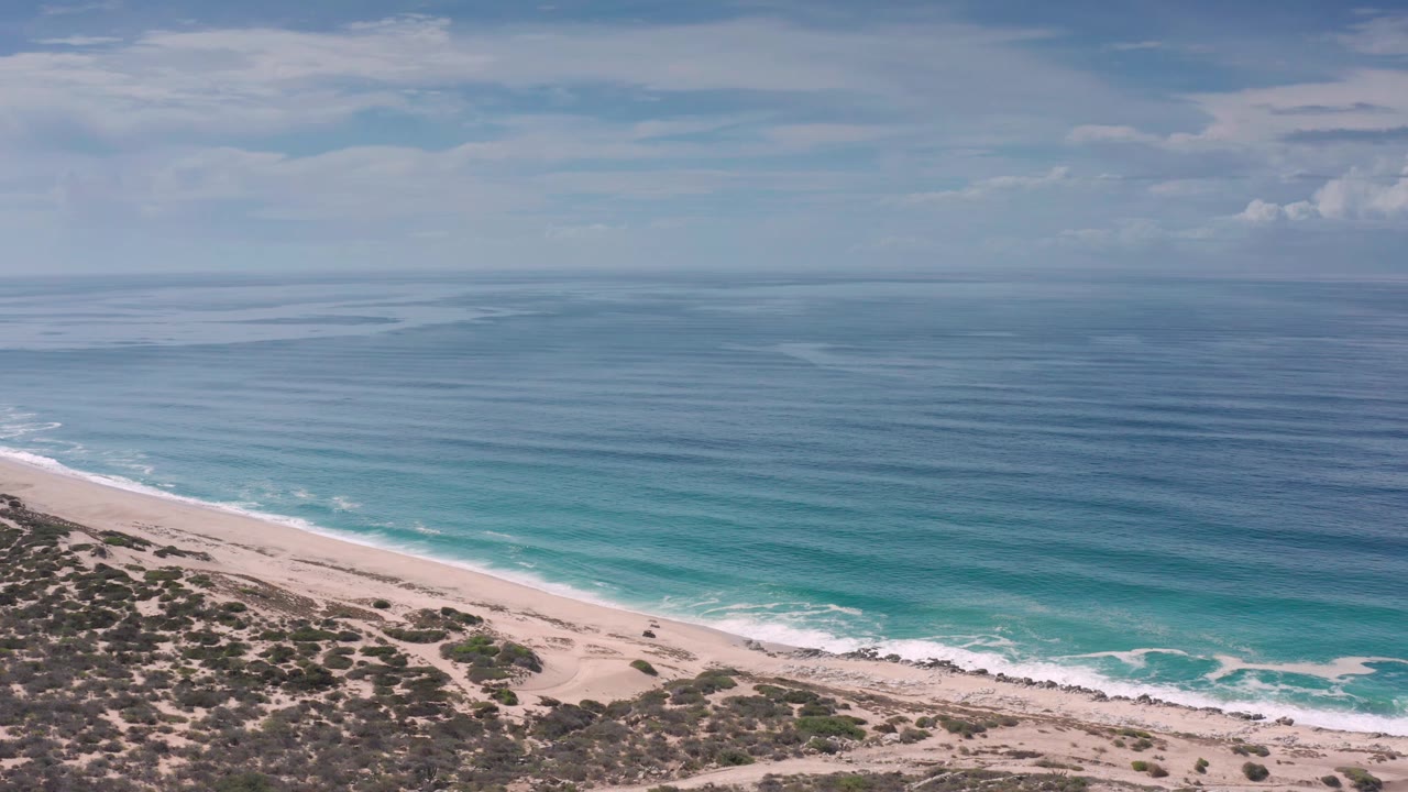 Los Cabos shoreline seen from above with a diagonal band of sand, white surf, blue-green water, darker ocean beyond, and no visible people.