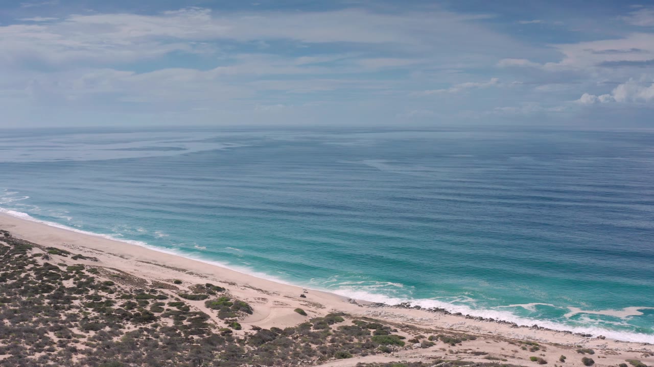 A wide aerial view in Los Cabos shows rocky scrub along the lower edge, a pale beach, low surf, a broad ocean surface, and no visible people.