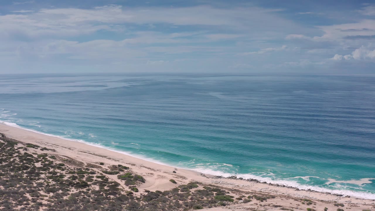 The Los Cabos beach edge sits below rolling white surf and layered blue water, with scrub-covered rocks at the bottom of frame and no visible people.
