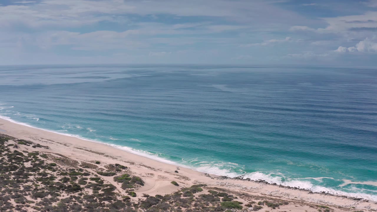 Los Cabos aerial coastline frame showing the beach diagonal across the lower image, low coastal vegetation, a thin white surf edge, layered blue-green water, and no visible people.