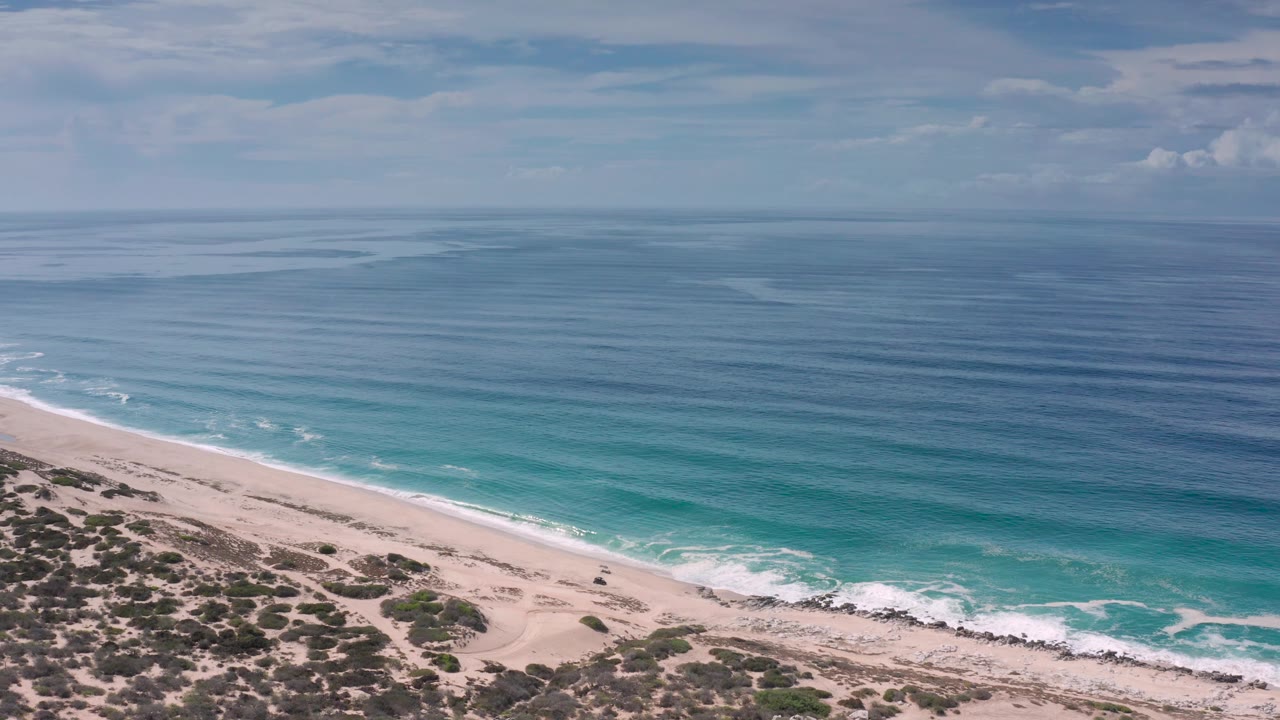 Wide aerial frame of the Los Cabos shoreline with scrubby dunes at the bottom, pale sand beach, white foam along the waterline, turquoise shallows, blue sea, soft clouds, and no visible people.