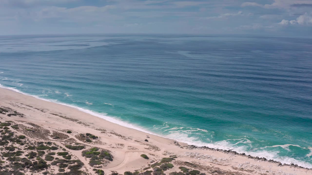 Opening aerial frame over a Los Cabos sandy beach with blue-green water, a diagonal surf line, dune scrub at the lower edge, and no visible people.