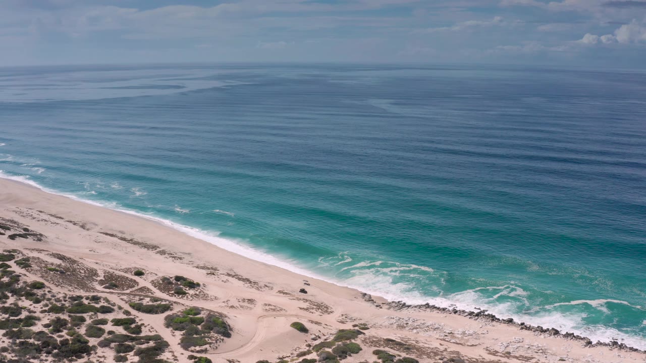 A wide aerial frame over the Los Cabos coast shows turquoise water near shore, pale sand below, scattered scrub and tracks on the beach, and no visible people.