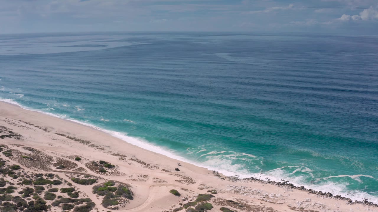 Mid-clip aerial view in Los Cabos with white surf washing onto the sandy shore, scrub patches in the lower frame, broad ocean above, and no visible people.