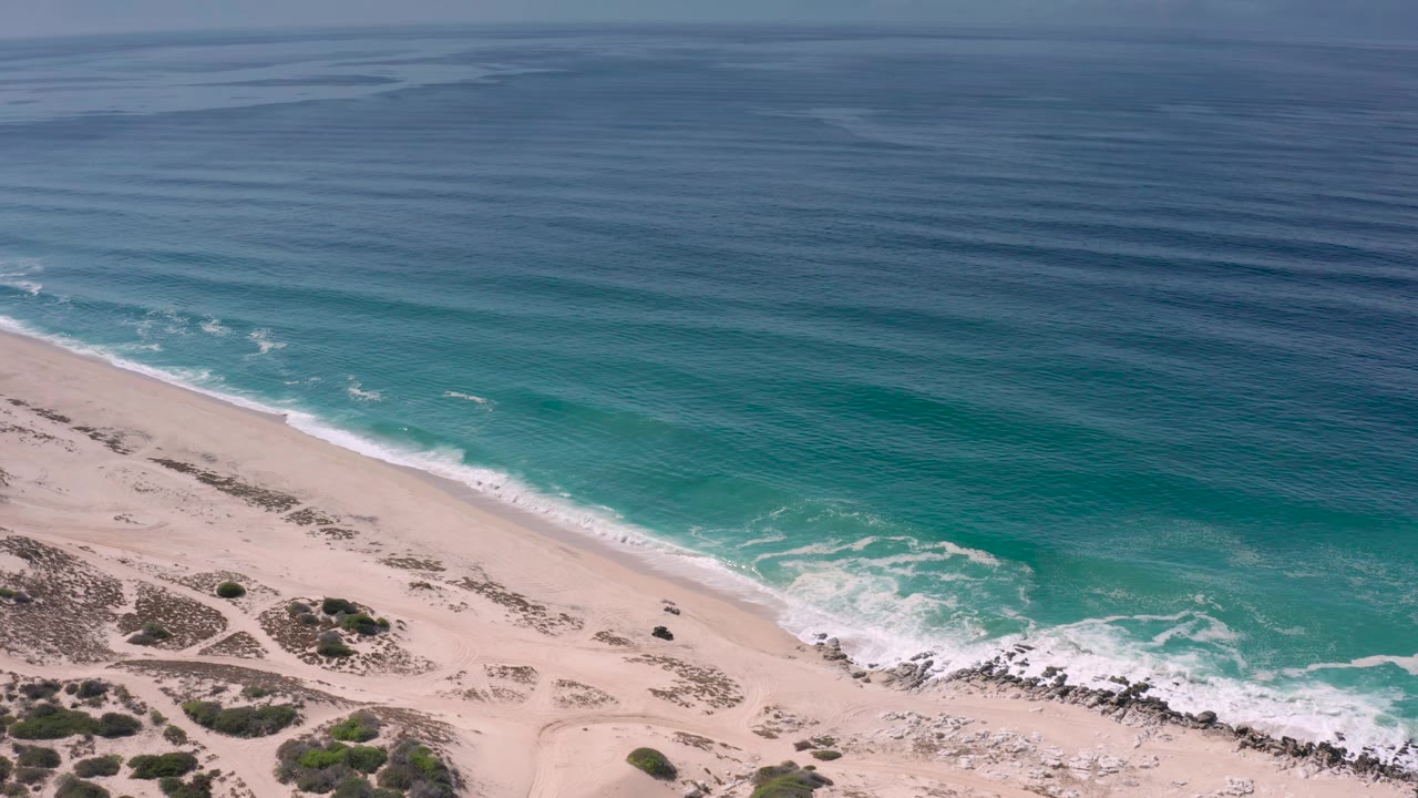 Aerial Los Cabos shoreline frame with blue-green water sliding toward the beach, a thin foam line, dune scrub, sand tracks, and no visible people.