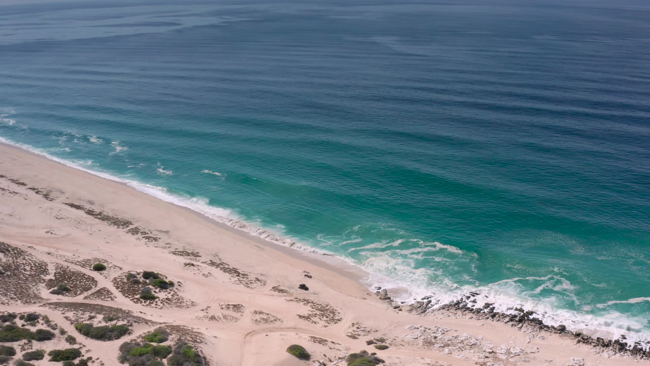 Oblique aerial frame from Los Cabos, Mexico with turquoise shore water, scattered white foam, a wide sand strip, dune vegetation, and no visible people.