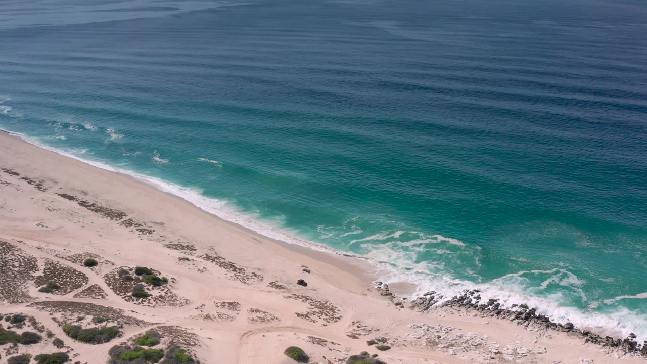 Los Cabos aerial beach frame showing soft wave foam along a diagonal shoreline, pale sand, dune scrub patches, faint vehicle or foot tracks, and no visible people.