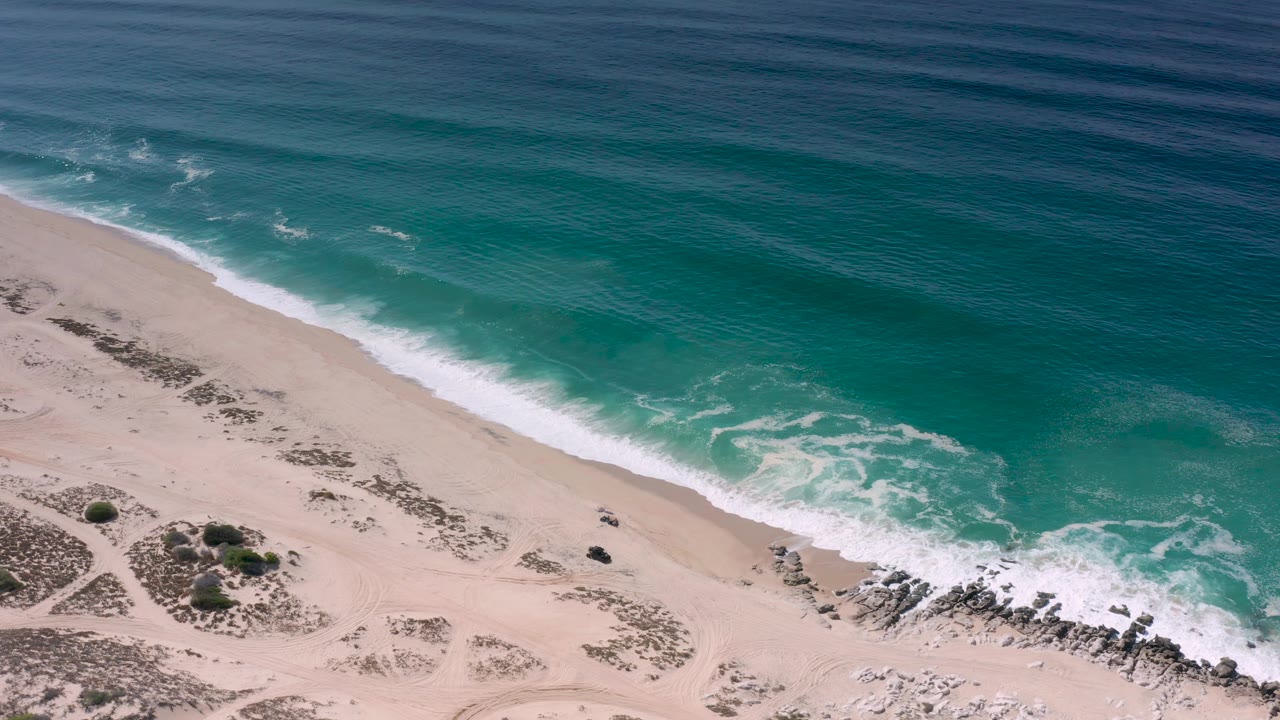 Aerial shoreline with blue ocean, white surf, pale sand, and rocks.