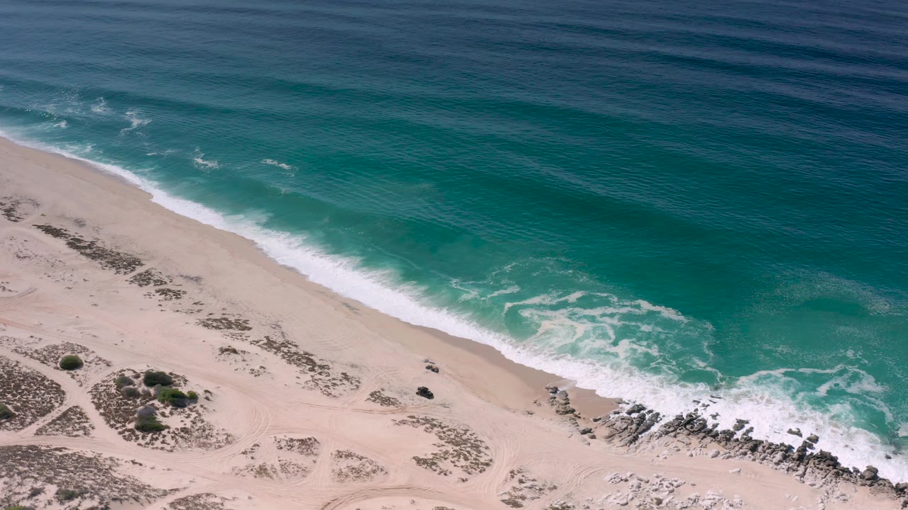 Aerial frame in Los Cabos, Mexico with turquoise water washing toward pale sand, low beach shrubs at lower left, rocks at lower right, and no visible people.