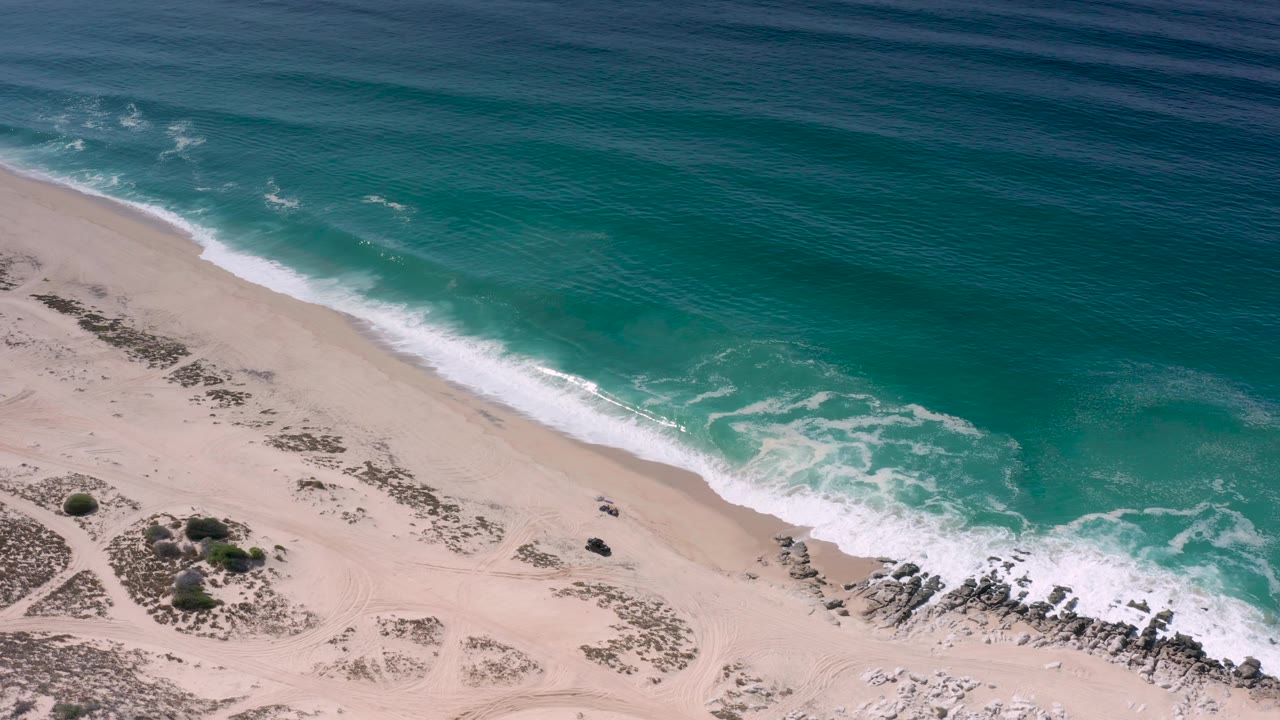 Aerial view in Los Cabos, Mexico showing blue-green ocean, a broad white foam band, pale sand, scattered dune vegetation, shoreline rocks, and no visible people.