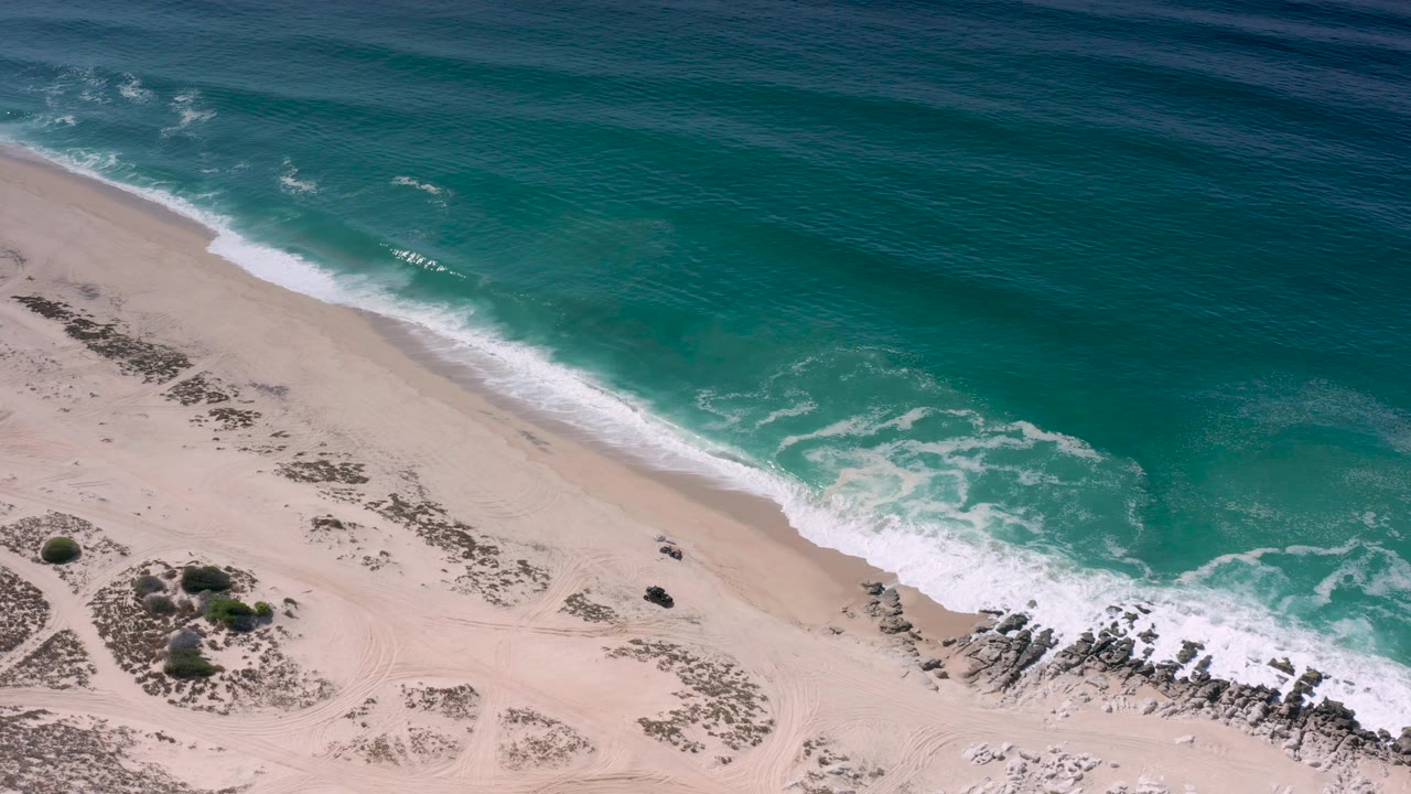 Aerial shot in Los Cabos, Mexico with darker blue water offshore, lighter turquoise water near the break, pale sand below, small shrubs, rocks, and no visible people.
