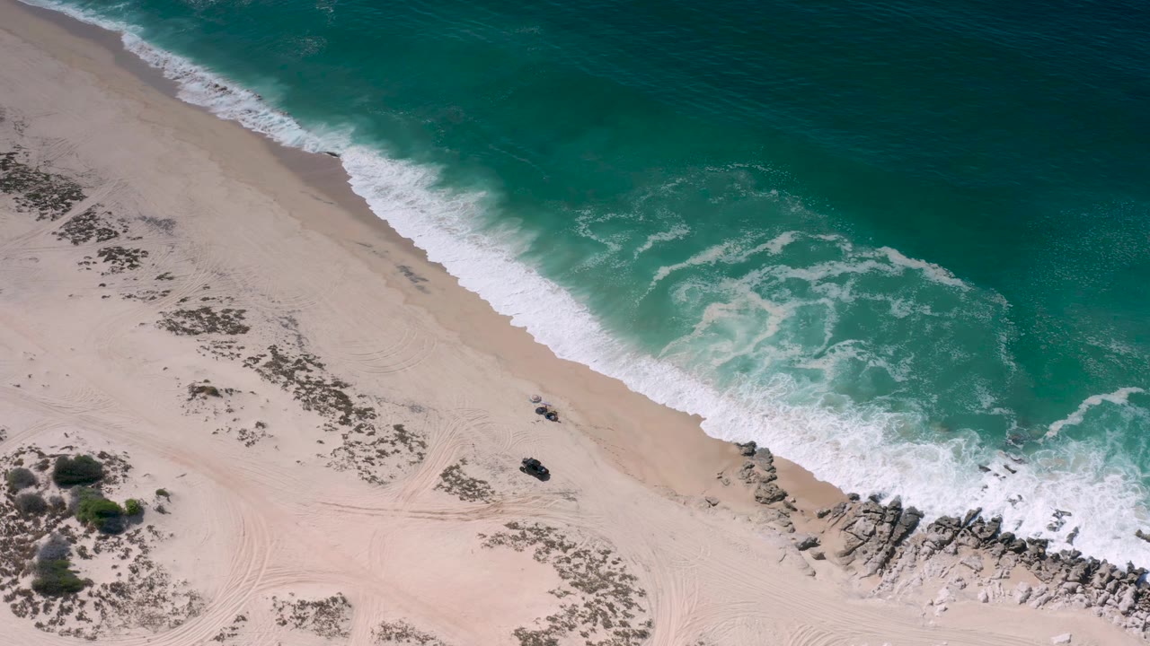 Overhead Los Cabos beach view with turquoise water, white foam, pale sand, scattered scrub, a rocky point, and no visible people at the start of the clip.