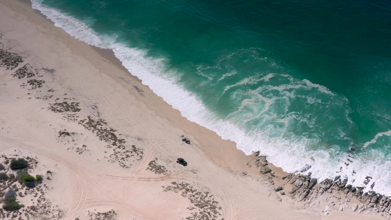 Blue-green water breaks onto the Los Cabos beach, with a curved foam edge, exposed sand, scrub clusters, rocks at the lower right, and no visible people.