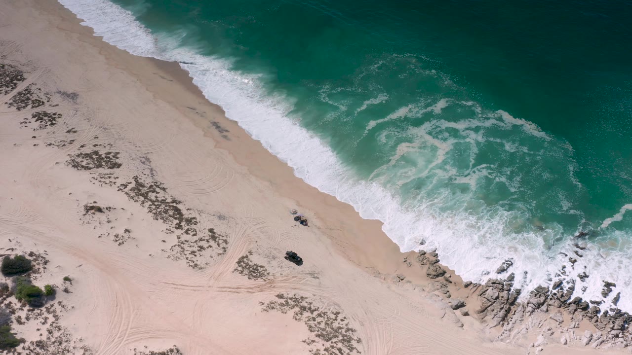 Overhead Los Cabos beach frame with foamy wave wash crossing pale sand, teal water above, scrub and track marks below, and no visible people.