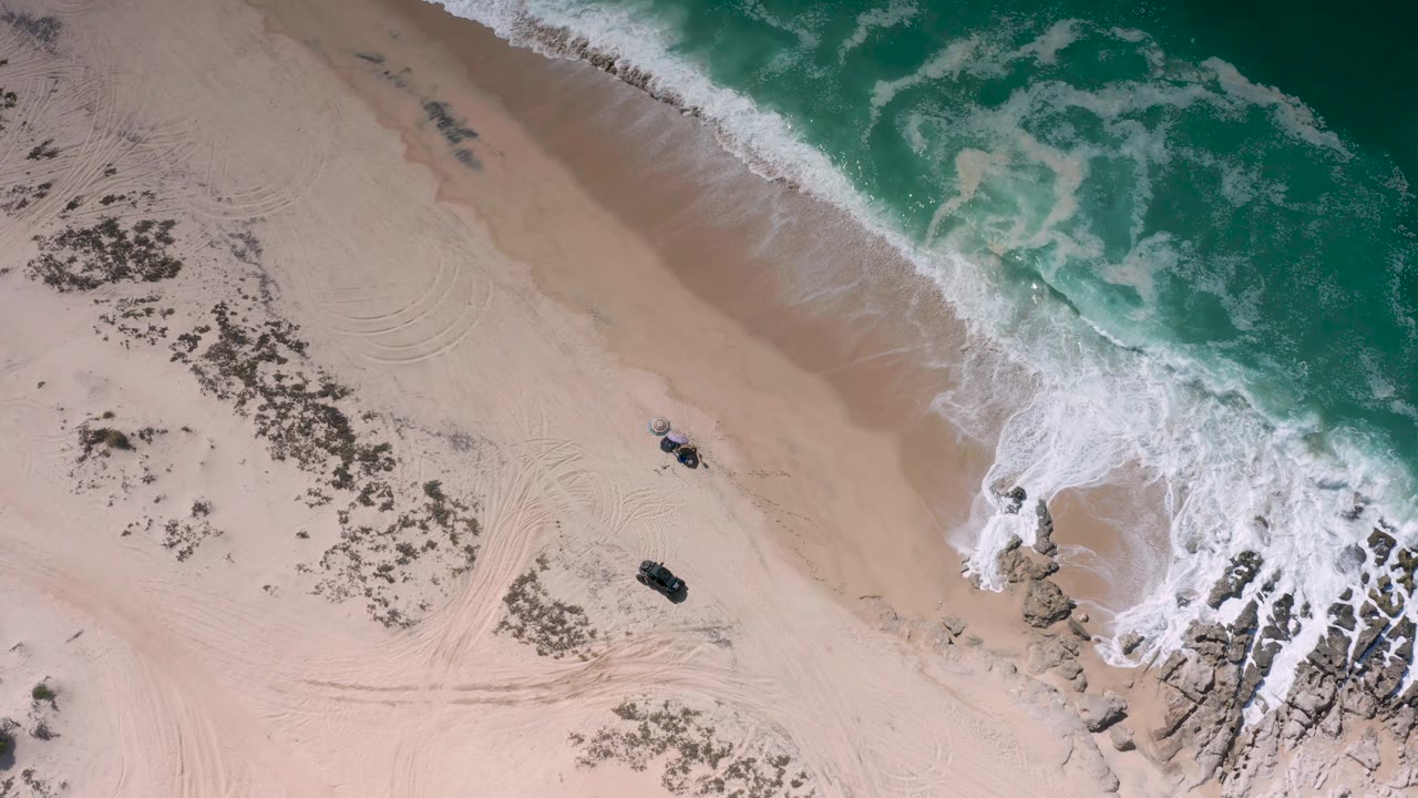 High aerial frame from Los Cabos, Mexico, with white foam curling over the upper right edge, pale sand below, curved tire tracks, rocks, and no visible people.