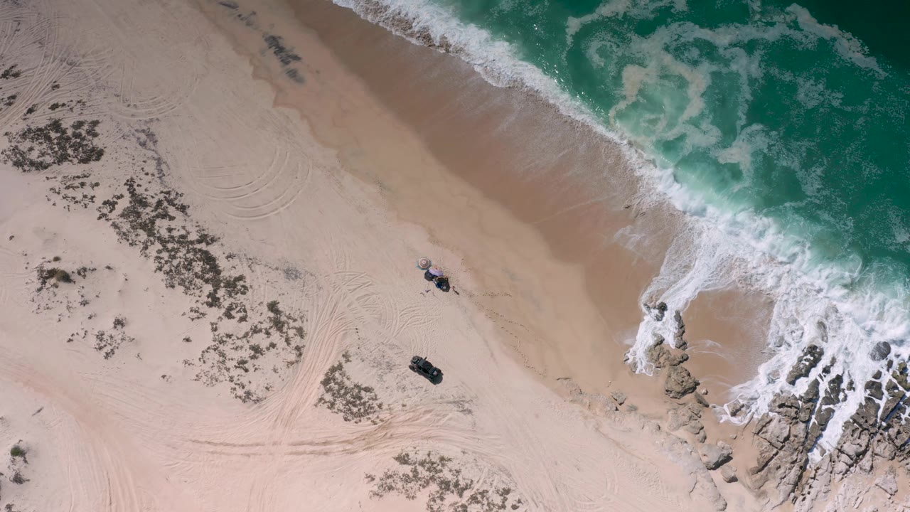 Top-down Los Cabos beach view with more surf entering from the upper right, a rock outcrop near the lower right shoreline, two small dark objects on the sand, and no visible people.