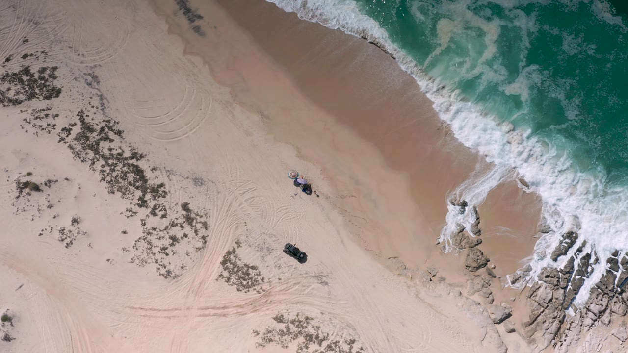 Drone view of beach with breaking surf, rocks, tire tracks, and scrub patches.
