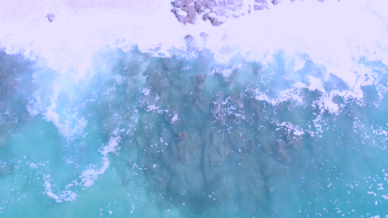Overhead aerial frame from Los Cabos, Mexico showing pale turquoise rough water washing over dark jagged rocks, with no visible people.