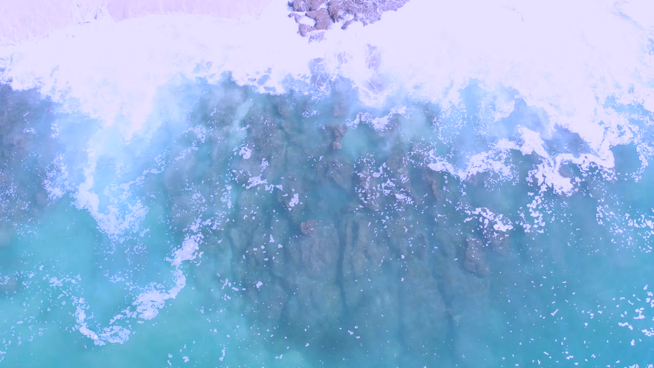 Overhead Los Cabos coastline detail with foamy surf breaking over jagged rocks in clear light blue water, with no visible people.