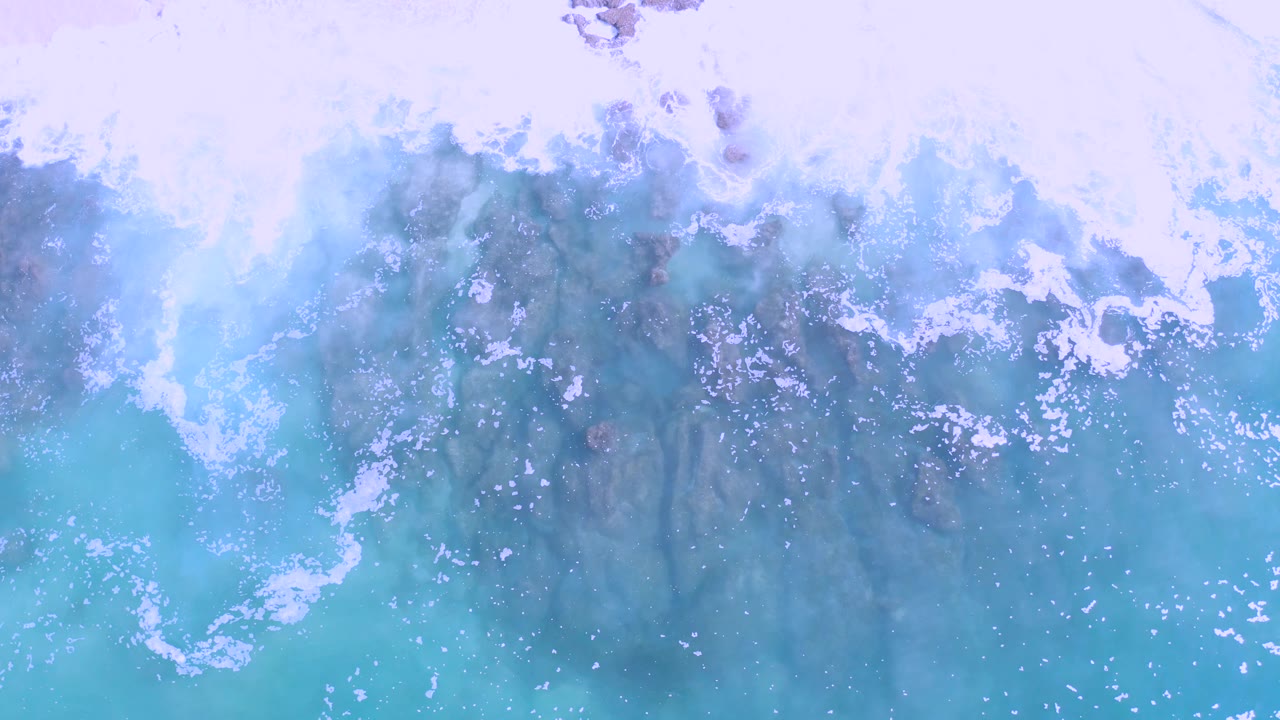 Mid-clip aerial view in Los Cabos, Mexico with rough turquoise surf, white wash, and darker rocks visible under the surface, with no visible people.