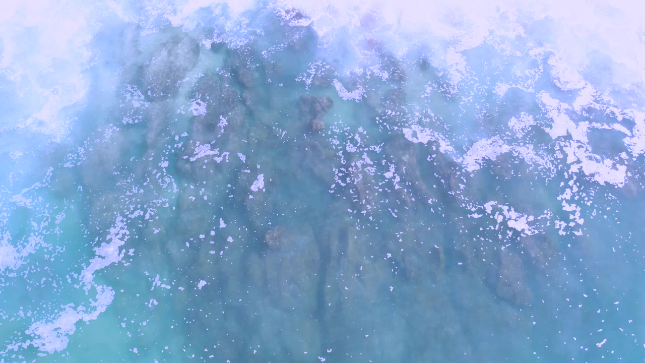 Overhead view in Los Cabos, Mexico of pale blue shallow water, scattered white sea foam, and darker submerged rocks, with no visible people.
