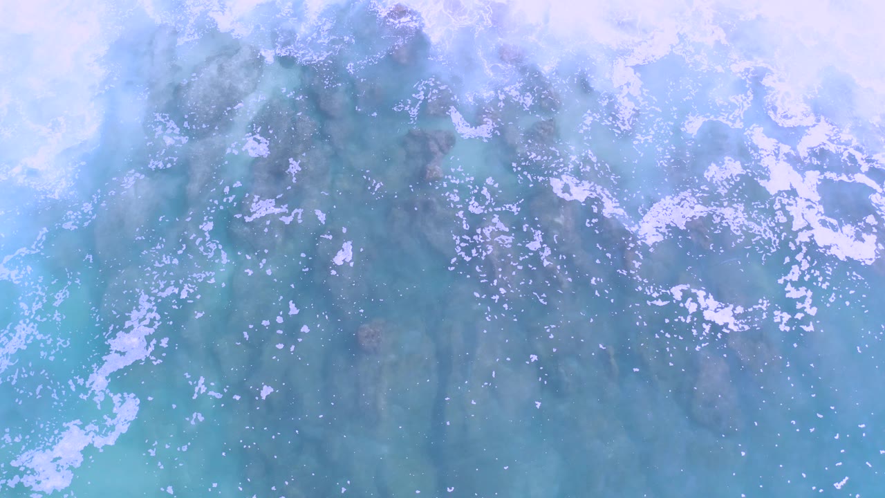 Top-down coastal water in Los Cabos, Mexico with a wider white foam streak over blue-green shallows and muted brown rocks below, with no visible people.