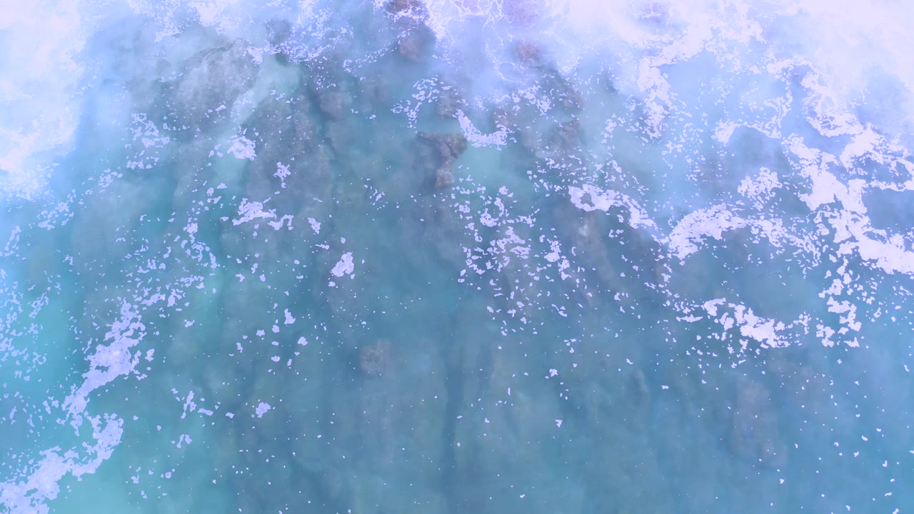 Overhead frame of soft blue surf, cloudy white foam, and shadowed underwater rock forms in Los Cabos, Mexico, with no visible people.