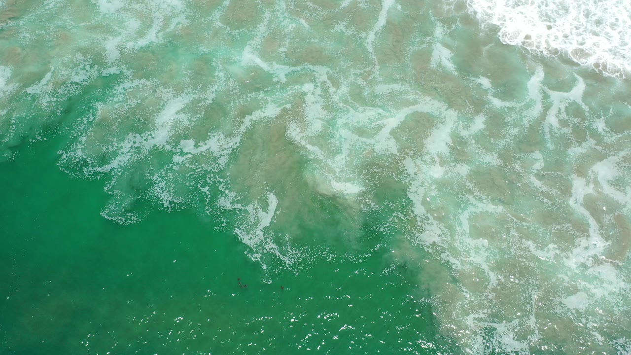 Overhead view of semi-translucent emerald water and white foam over shallow sand in Los Cabos, Mexico, with no visible people.
