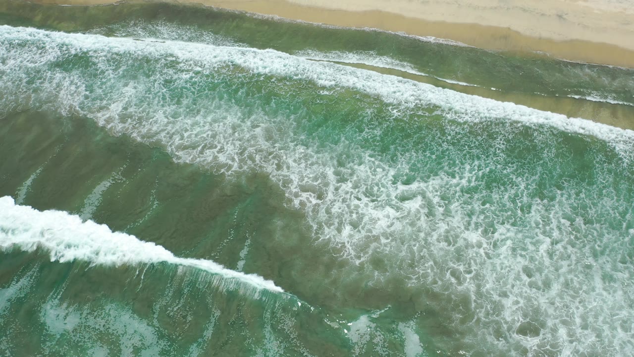 Aerial frame of a long white breaker cutting diagonally through green ocean water near the Los Cabos shoreline, with no visible people.