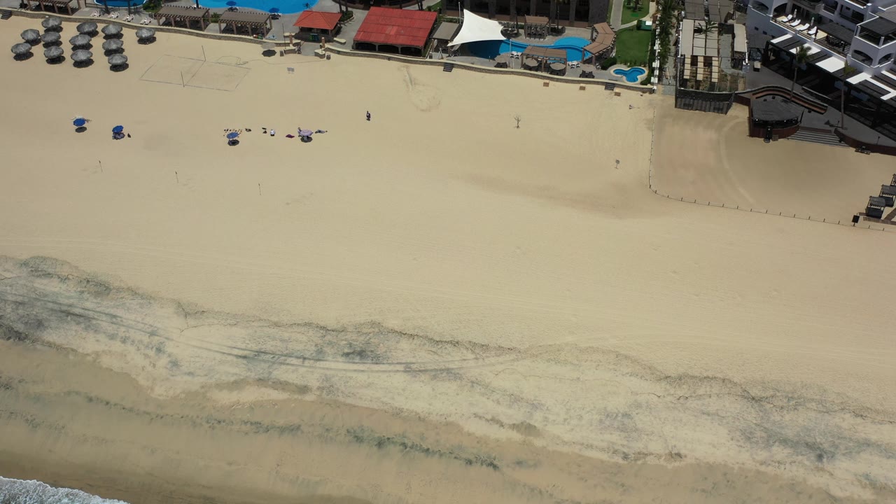 High aerial frame over Los Cabos beach with blue resort pools and red-roofed structures at the top, open sand through the center, and uneven surf marks along the lower edge, with no clearly visible people.