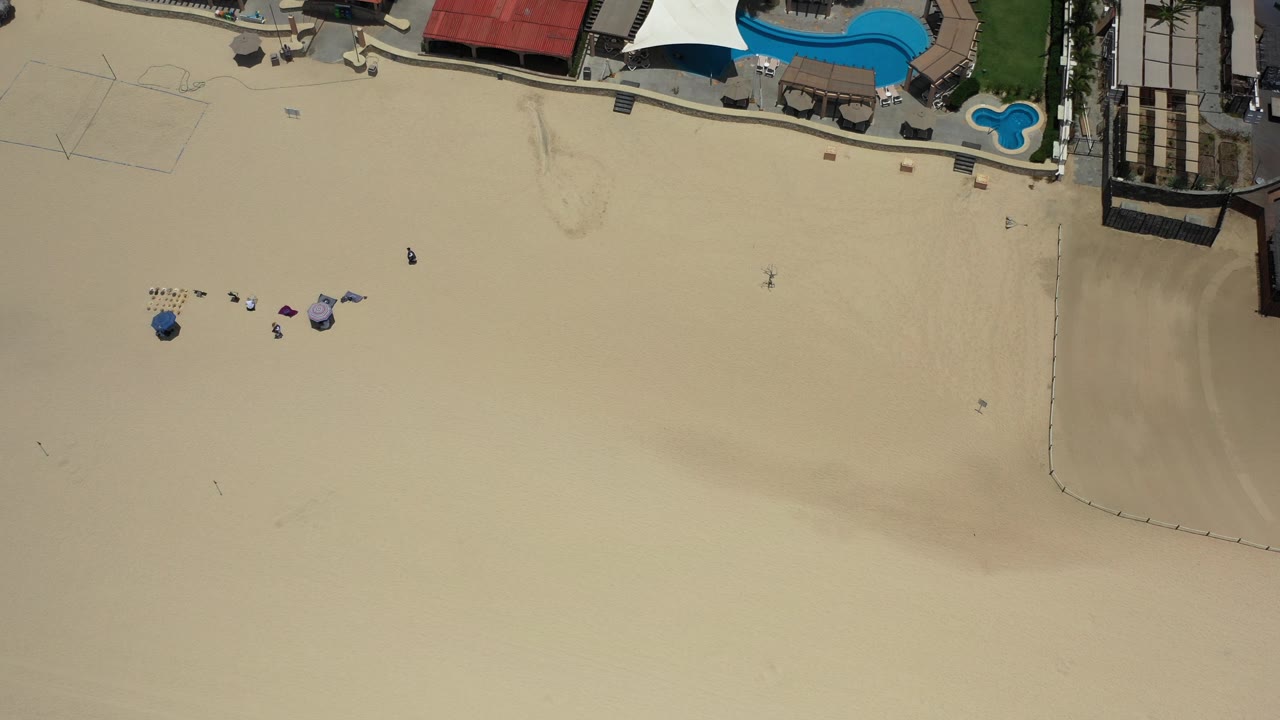 Top-down aerial frame in Los Cabos, Mexico, showing broad pale sand, small visible people, grouped umbrellas and chairs, and blue resort pools along the upper edge.