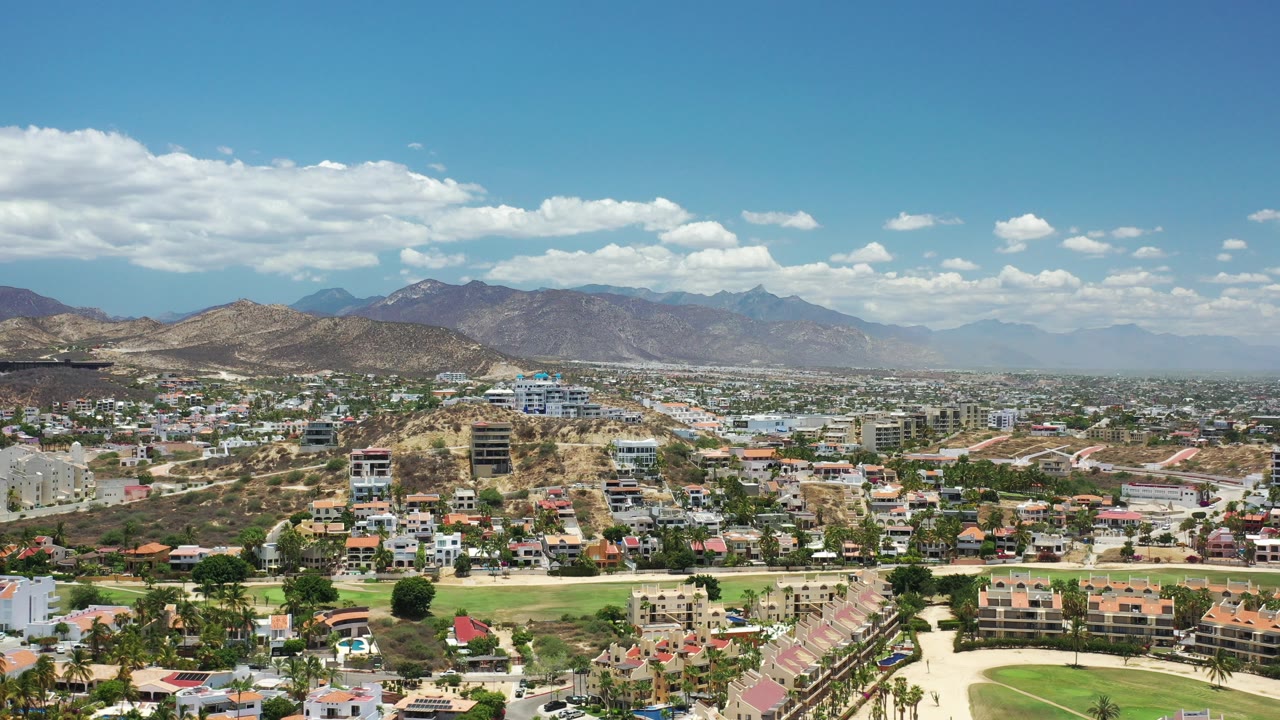 Opening aerial frame over Los Cabos, Mexico, showing hillside homes, roads, dry ridges, distant mountains, scattered clouds, and no visible people.