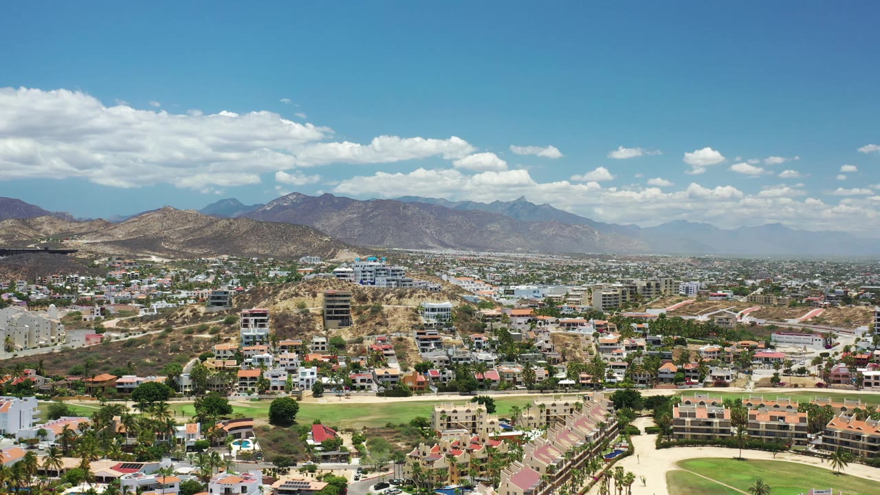Early aerial view of Los Cabos, Mexico, with dense hillside housing in the lower frame, tan ridgelines at left, mountains across the horizon, and no visible people.