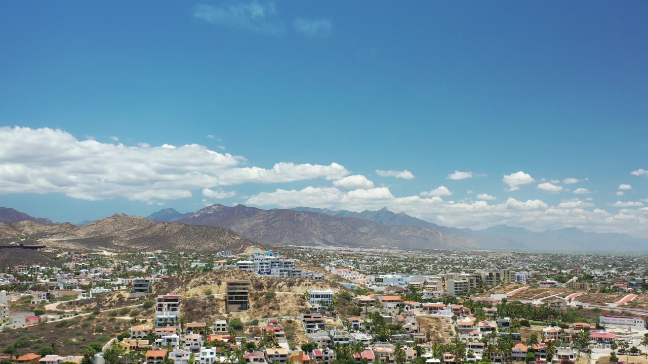 Wide aerial view of a city below dry hills and mountains under a blue sky.