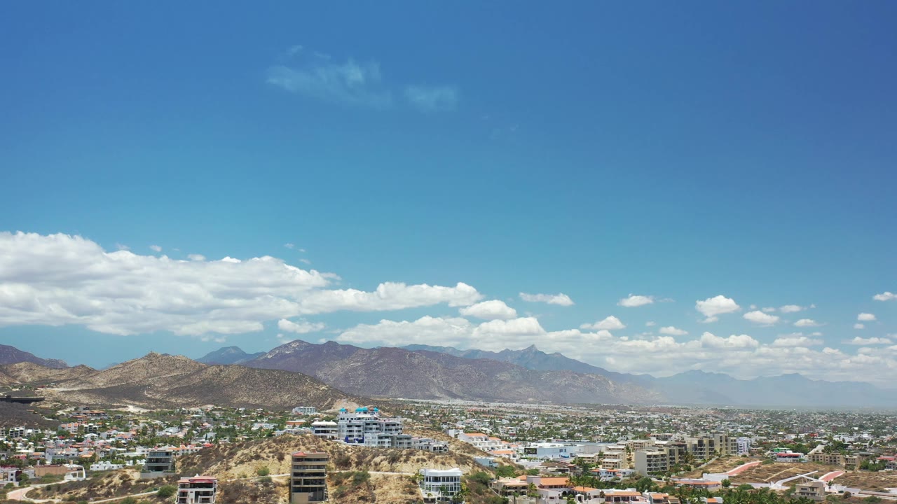 Los Cabos city rooftops and roads sit beneath a broad blue sky, with dry hills and a layered mountain range across the horizon and no visible people.