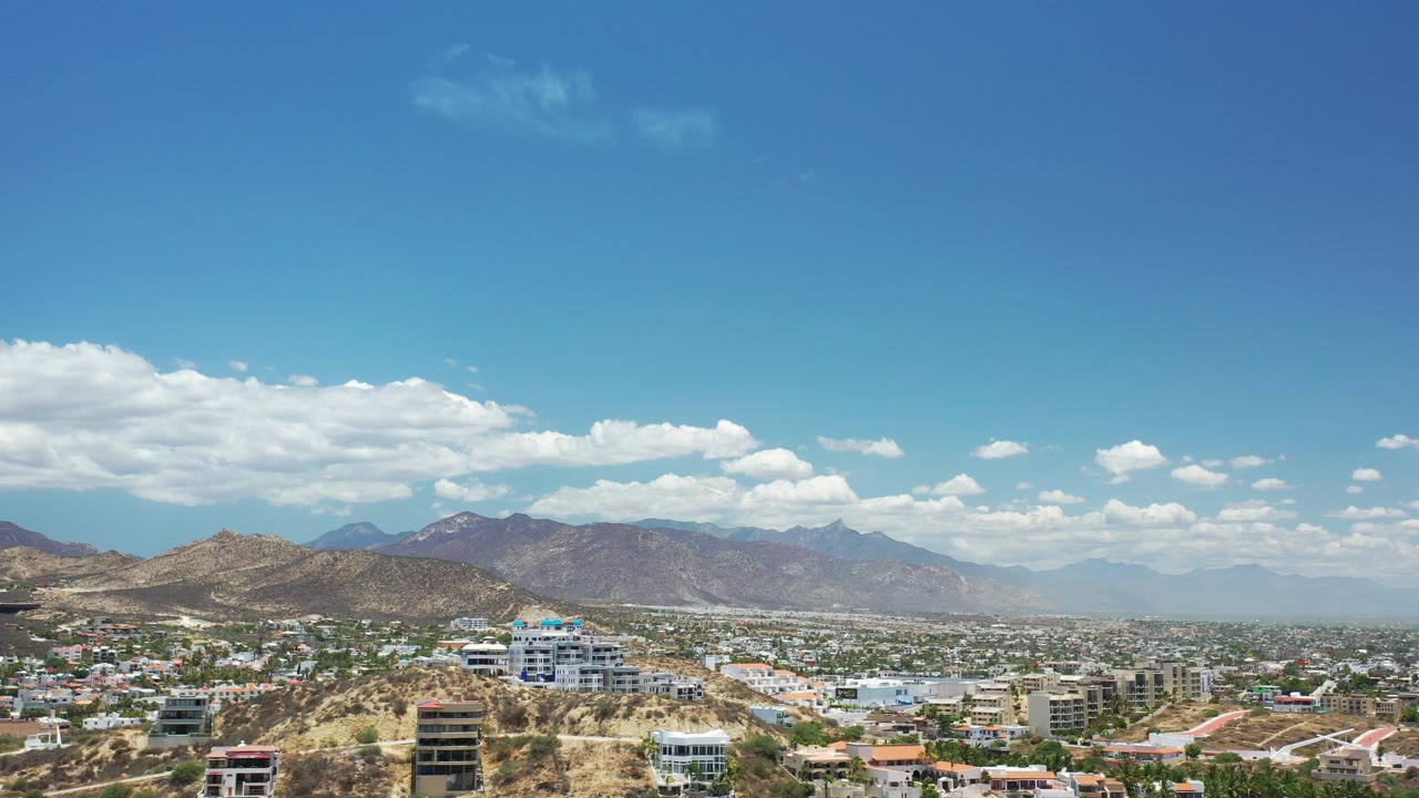 Opening frame of Los Cabos, Mexico from a high vantage point, with hillside homes in front, city blocks across the valley, mountains beyond, scattered clouds, and no visible people.