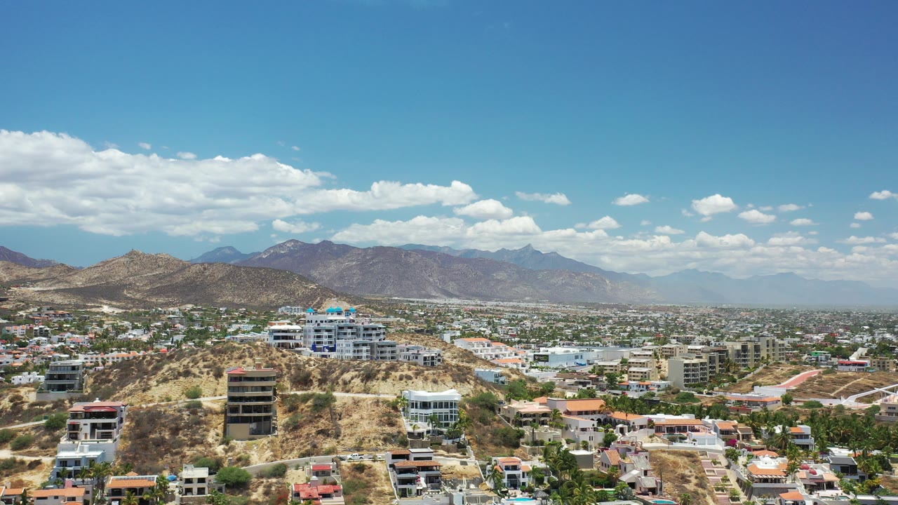 Wide aerial view of hillside homes, town buildings, desert terrain, mountains, and blue sky.