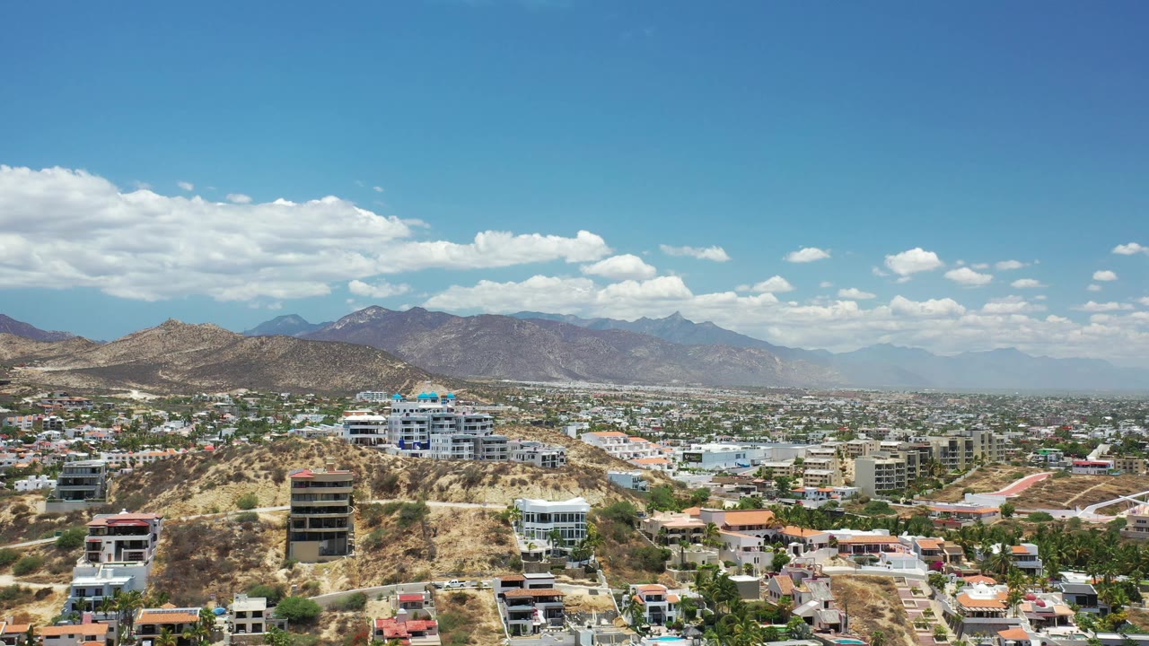 Aerial frame in Los Cabos, Mexico showing hillside residences and a denser residential area below a mountain range, with bright midday light and no visible people.