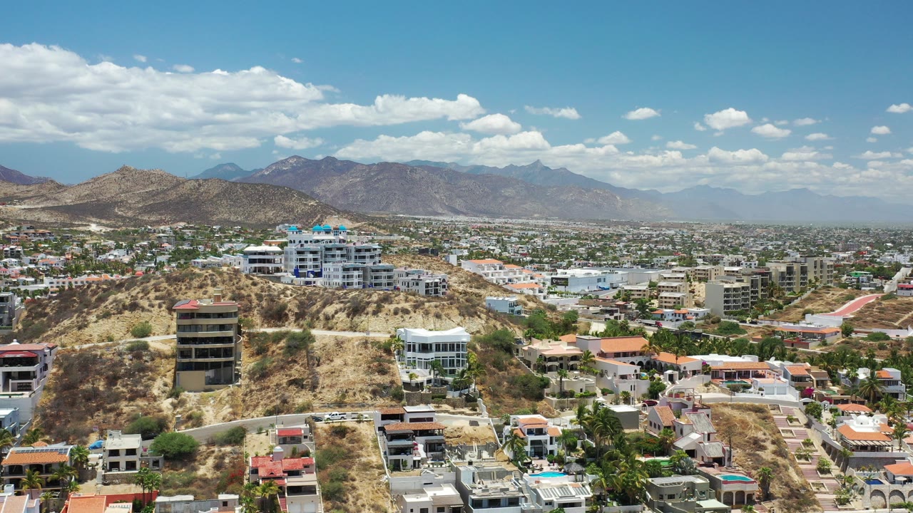 Wide aerial view of hillside homes, roads, and distant mountains under clouds.