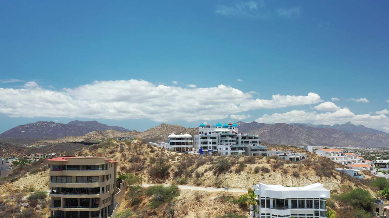 Wide elevated view in Los Cabos, Mexico of modern homes on dry rocky hills with distant mountains, bright clouds, and no visible people.